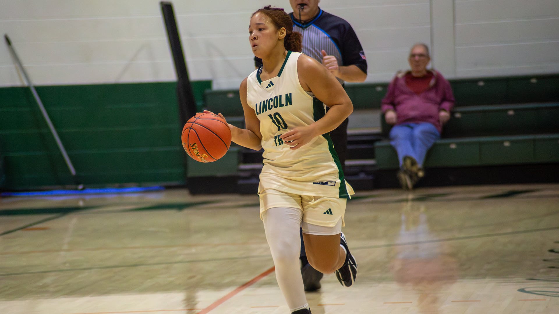 Khamryn Hopkins of Lincoln Trail College dribbles the basketball up the court during a game, wearing a white Statesmen uniform with number 10. A referee follows the play behind her as spectators watch from the bleachers in the background.